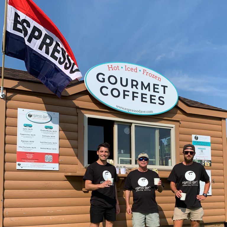 3 baristas standing with coffee cups in front of a log cabin coffee concession.