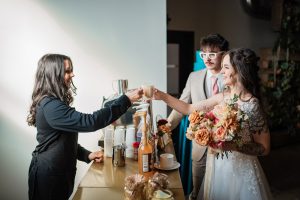 Female barista in black apron handing bride a cappuccino in a glass mug while groom watches beside Espresso Dave Coffee Catering espresso bar in at Boston wedding reception. Capture the North Photography