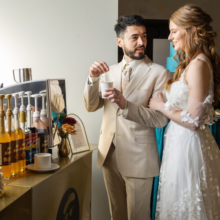 Groom and Bride smiling at each other in front of a gold espresso cart by Espresso Dave while groom stirs his latte in a white paper cup