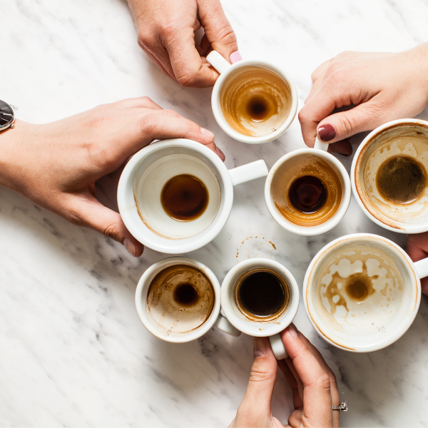 Image for a blog post on How Often Should You Clean Your Coffee Mug. It's a View from above of hands grasping their empty, dirty, white, coffee cups of various sizes on a marble table